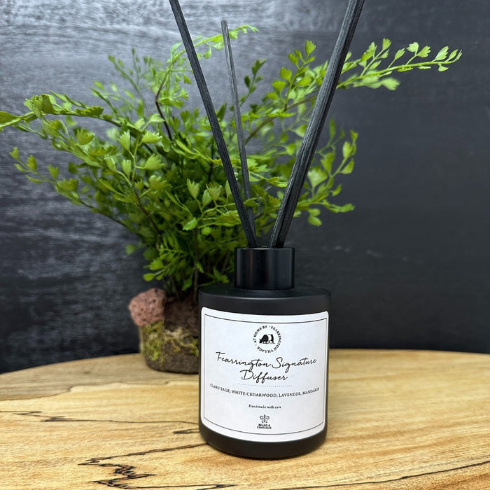 Housed in a sleek black bottle with five slender black reeds, the FEARRINGTON SIGNATURE DIFFUSER from the FEARRINGTON SIGNATURE COLLECTION by FEARRINGTON VILLAGE - HOME DECOR features a white label displaying "Fragrance Signature Diffuser" and scent notes of cedarwood, lavender, and amber. Inspired by the tranquil allure of Fearrington Village, this sophisticated reed diffuser is ideal for anyone who appreciates the calming atmosphere created by a lavender candle.