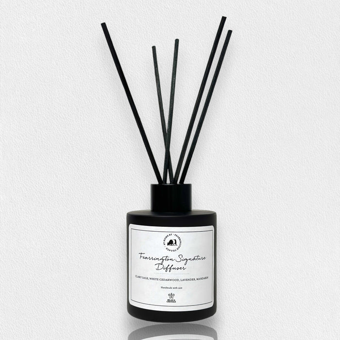 Housed in a sleek black bottle with five slender black reeds, the FEARRINGTON SIGNATURE DIFFUSER from the FEARRINGTON SIGNATURE COLLECTION by FEARRINGTON VILLAGE - HOME DECOR features a white label displaying "Fragrance Signature Diffuser" and scent notes of cedarwood, lavender, and amber. Inspired by the tranquil allure of Fearrington Village, this sophisticated reed diffuser is ideal for anyone who appreciates the calming atmosphere created by a lavender candle.