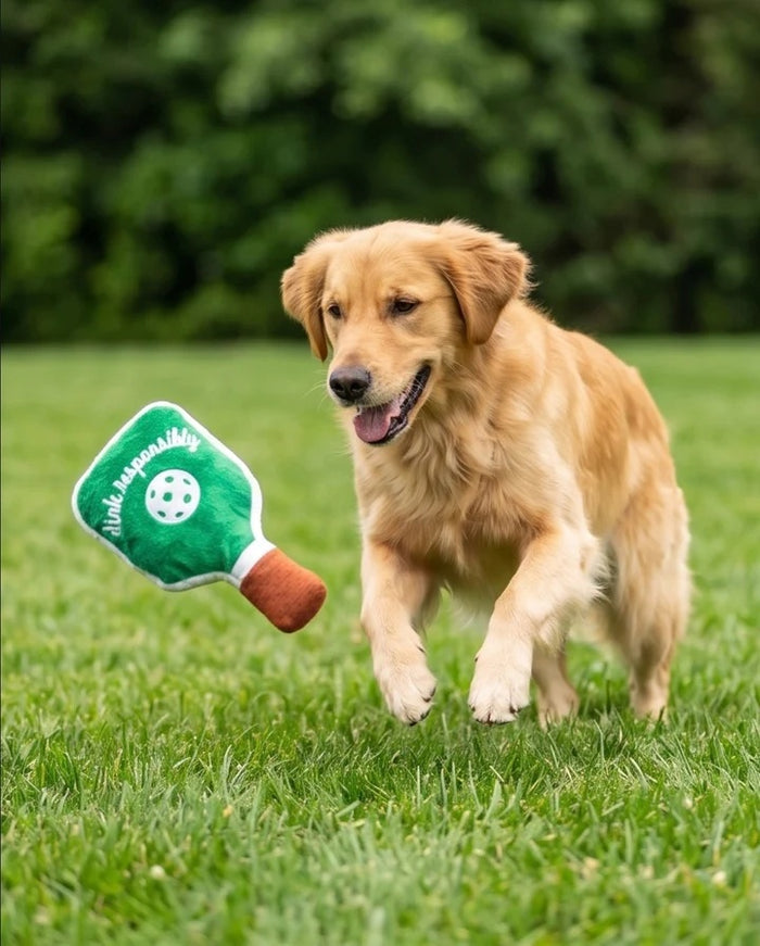 The HAUTE DIGGITY DOG GREEN STRIPED PICKLEBALL PADDLE DOG TOY is a plush squeaky dog toy shaped like a green and white striped pickleball paddle with a green ball in the center and "just dink it." embroidered in pink on the front, making it the perfect pickleball dog toy for playful pups.