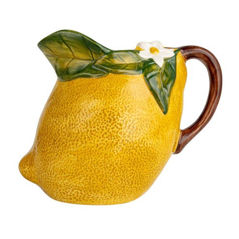 The HAND-PAINTED STONEWARE LEMON-SHAPED PITCHER by CREATIVE COOP, featuring a ceramic lemon form adorned with delicate floral and leafy details near the handle, is displayed beside a folded cloth, a halved grapefruit, and a yellow dish on a light background.