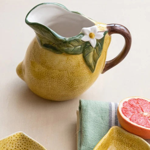 The HAND-PAINTED STONEWARE LEMON-SHAPED PITCHER by CREATIVE COOP is a ceramic piece crafted in the form of a vibrant yellow lemon, complete with a green leaf accent, delicate white flower, and brown handle.