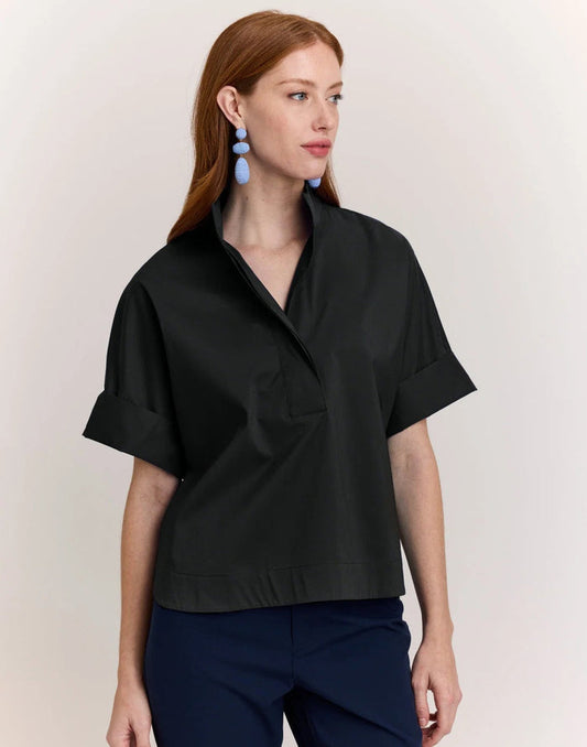 A woman with long red hair wears the HINSON WU Cara Elbow Sleeve Shirt in Stretch Polished Cotton Black—an oversized, loose-fit blouse by HINSON WU—paired with navy pants and dangling blue earrings, standing against a plain light background.