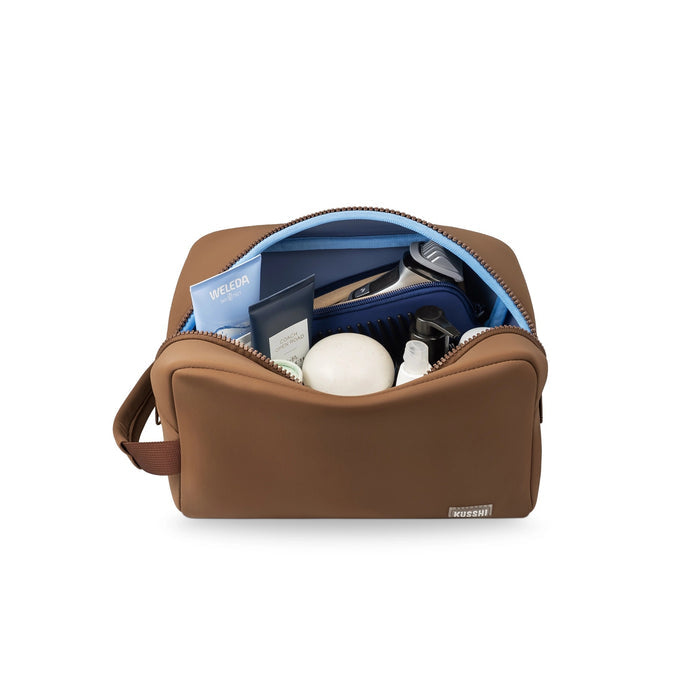 The KUSSHI Neoprene Dopp Kit in Chocolate is a brown rectangular unisex toiletry bag with a top zipper, side handle, and small white "KUSSHI" logo—ideal as a travel organizer for your essentials.