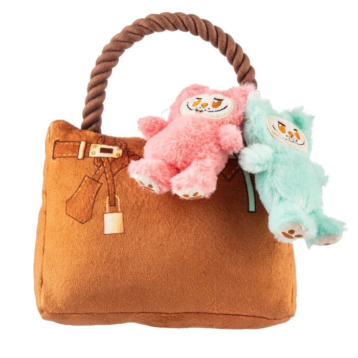 A French Bulldog sits indoors in a bright, cozy room, holding the HAUTE DIGGITY DOG LABOWWOW HANDBAG DOG TOY—a durable plush handbag adorned with pink and blue teddy bear keychains—in its mouth.