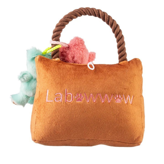 A French Bulldog sits indoors in a bright, cozy room, holding the HAUTE DIGGITY DOG LABOWWOW HANDBAG DOG TOY—a durable plush handbag adorned with pink and blue teddy bear keychains—in its mouth.