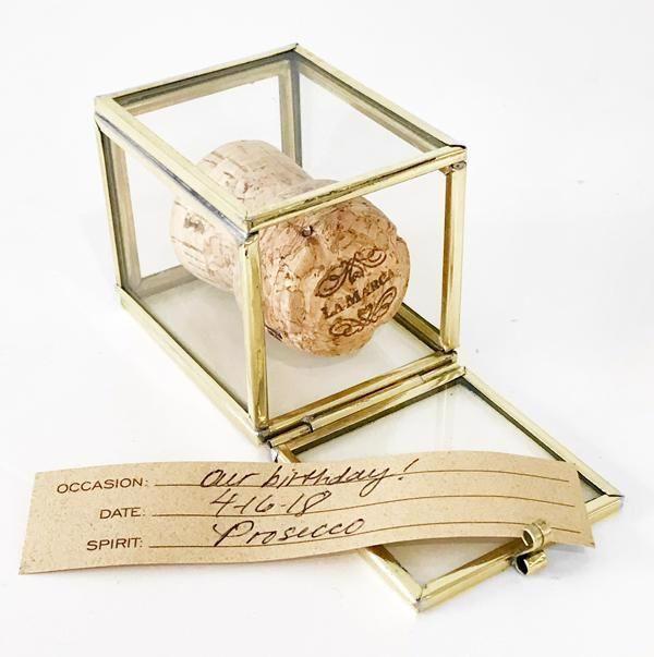 The JM PIERS LARGE CORK SAFE displays a large prosecco cork in a glass and brass box with a handwritten tag: Occasion: Our birthday! Date: 4.6.18 Spirit: Prosecco – a stylish keepsake for commemorating special moments.