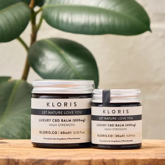 Two jars of KLORIS CBD balm on a wooden surface with green leaves in the background