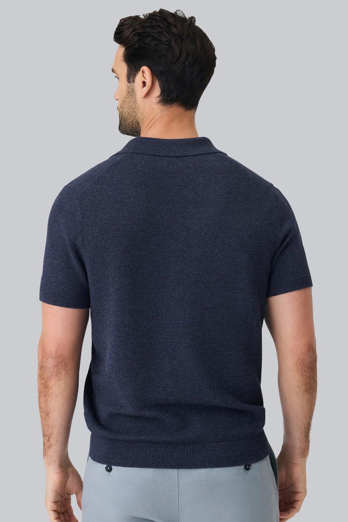 A close-up of a person’s arm and torso wearing the MACK WELDON Tech Linen Johnny Collar Polo Sweater in Navy Heather, paired with light gray pants featuring a buttoned pocket.