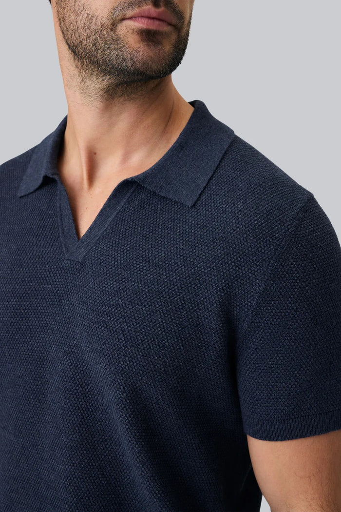 A close-up of a person’s arm and torso wearing the MACK WELDON Tech Linen Johnny Collar Polo Sweater in Navy Heather, paired with light gray pants featuring a buttoned pocket.