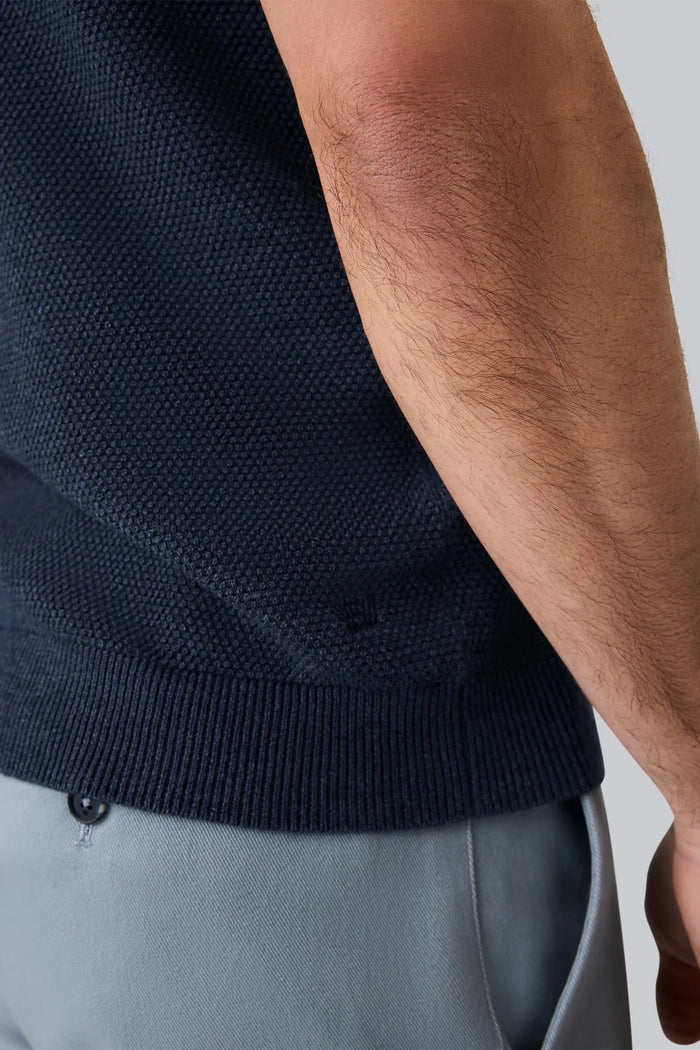 A man with short dark hair and a trimmed beard models the MACK WELDON Tech Linen Johnny Collar Polo Sweater in Navy Heather with light gray pants, standing against a plain white background.