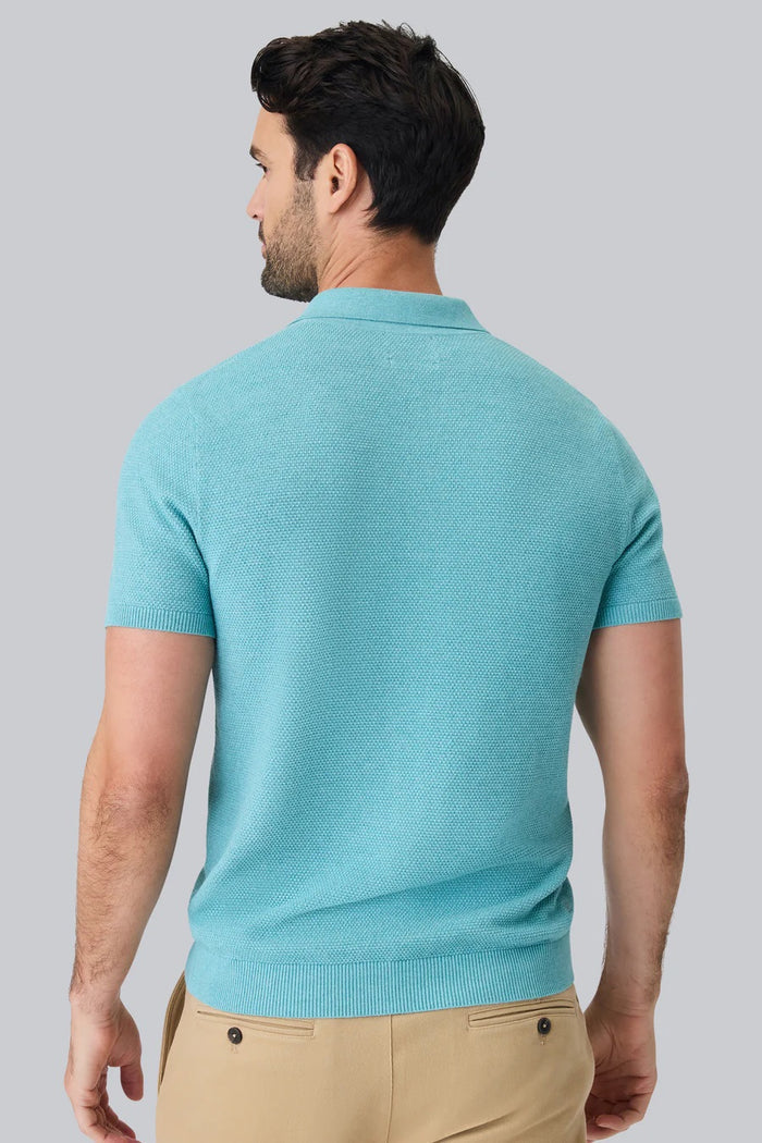 A man with short dark hair and a beard wears the MACK WELDON TECH LINEN SWEATER POLO in Deep Lagoon Heather, made with COOLMAX technology, and beige pants. He looks down, left arm bent, posed against a plain light gray background.