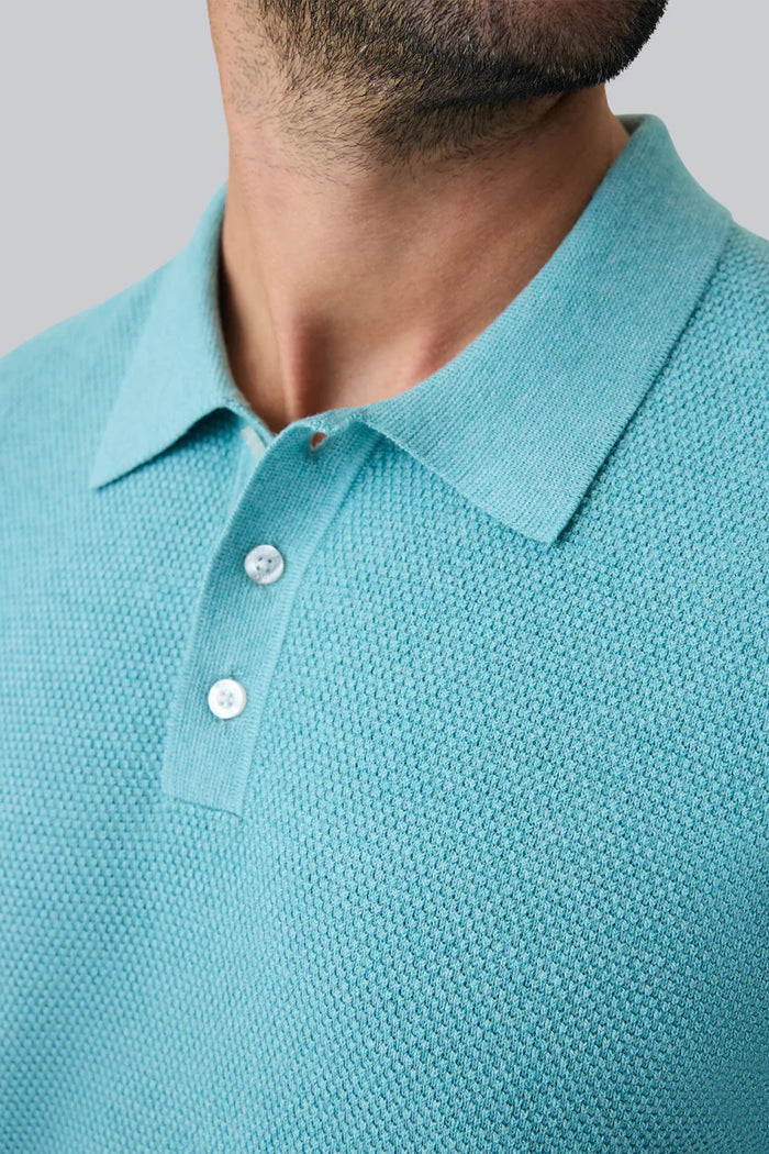A man with short dark hair and a beard wears the MACK WELDON TECH LINEN SWEATER POLO in Deep Lagoon Heather, made with COOLMAX technology, and beige pants. He looks down, left arm bent, posed against a plain light gray background.