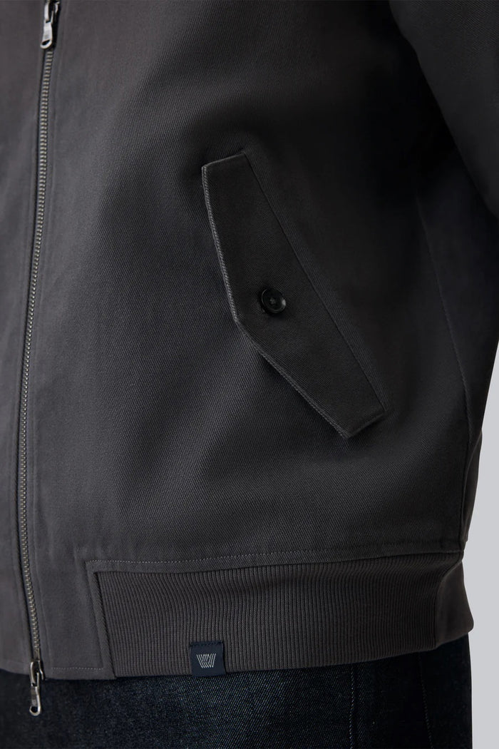 The MACK WELDON Twill Harrington Jacket in Asphalt is a dark gray cotton cavalry twill zip-up with a stand collar, two angled button-flap pockets, ribbed cuffs and hem, offering an English jacket style. Shown on a black background.