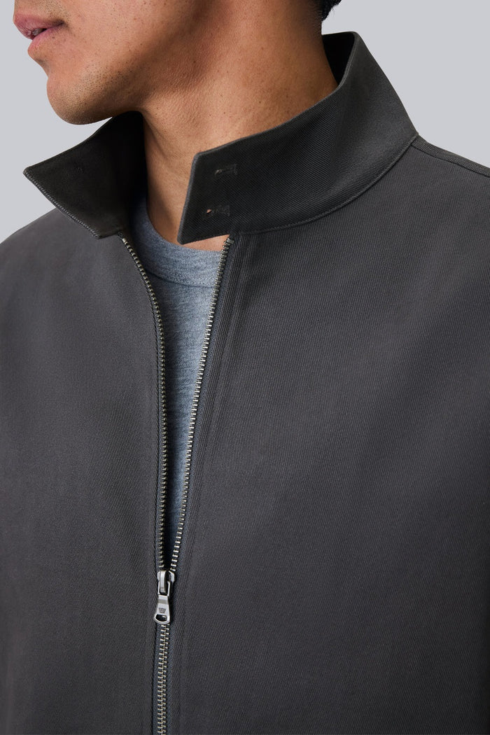 Close-up of a person wearing the MACK WELDON Twill Harrington Jacket in Shroom, featuring a zip front and buttoned flap pocket. Their arm is relaxed by their side and dark denim jeans are partially visible.
