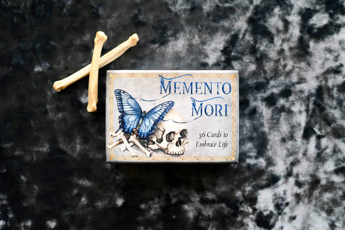Memento Mori Cards box with butterfly and skull design on dark textured background.
