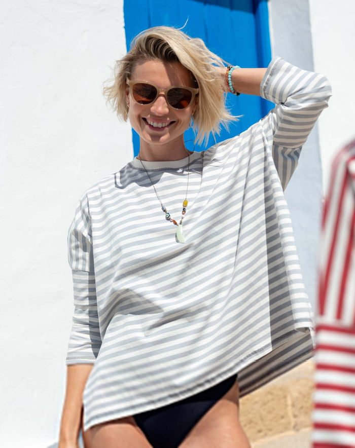 A smiling person with short blonde hair and sunglasses is wearing the MERSEA - CATALINA JERSEY TEE IN SLATE BLUE by MERSEA, along with a necklace and bracelets, standing outdoors in front of a blue door and white wall.