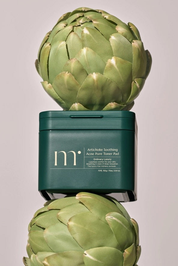A green container of "MOLVANY - Artichoke Soothing Affect Pore Toner Pads" by HONG CHEON USA, INC is balanced between two whole artichokes on a neutral background. Its minimalist design emphasizes its non-comedogenic nature, ideal for acne-prone skin.