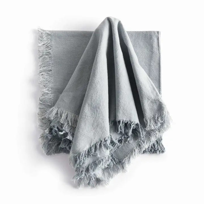 A table setting features a white plate, bowl, and textured placemat. The NAPA HOME AND GARDEN - AURA DINNER NAPKIN IN LIGHT BLUE is knotted atop the plates, with silverware arranged neatly alongside.