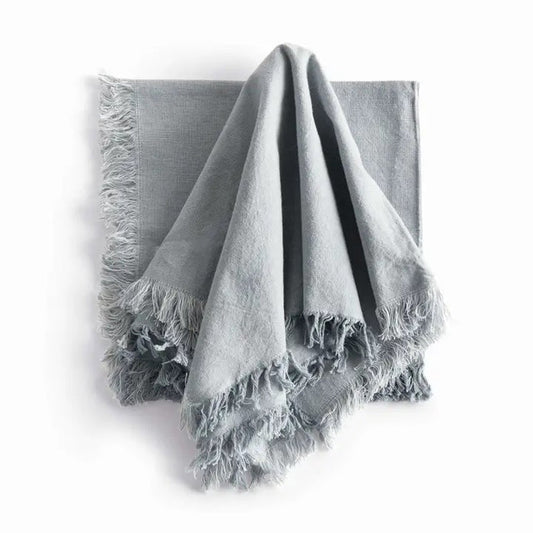 A table setting features a white plate, bowl, and textured placemat. The NAPA HOME AND GARDEN - AURA DINNER NAPKIN IN LIGHT BLUE is knotted atop the plates, with silverware arranged neatly alongside.