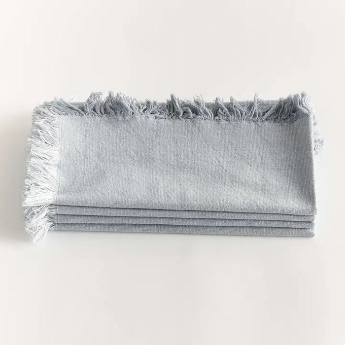 A table setting features a white plate, bowl, and textured placemat. The NAPA HOME AND GARDEN - AURA DINNER NAPKIN IN LIGHT BLUE is knotted atop the plates, with silverware arranged neatly alongside.