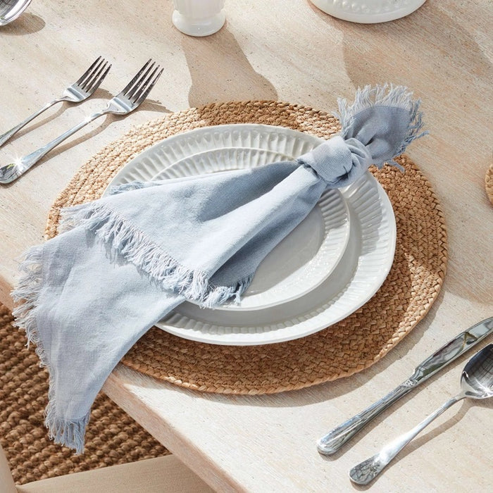 A light gray, fringed throw blanket is draped on a flat surface, highlighting its softness and decorative edges—perfectly paired with the NAPA HOME AND GARDEN AURA Dinner Napkin in Light Blue for an inviting table setting.