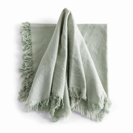 A stack of NAPA HOME AND GARDEN - AURA DINNER NAPKIN IN LIGHT GREEN, featuring fringed edges, is folded and placed on a white surface.