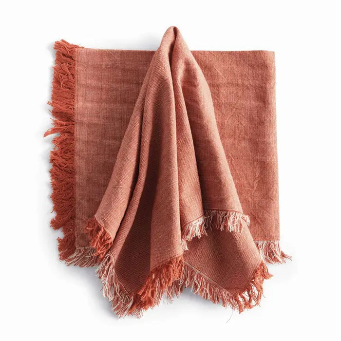 A table setting features white plates on woven placemats, a NAPA HOME AND GARDEN Aura Dinner Napkin in Rust with fringed edges knotted on top, and neatly arranged silverware on a light table.