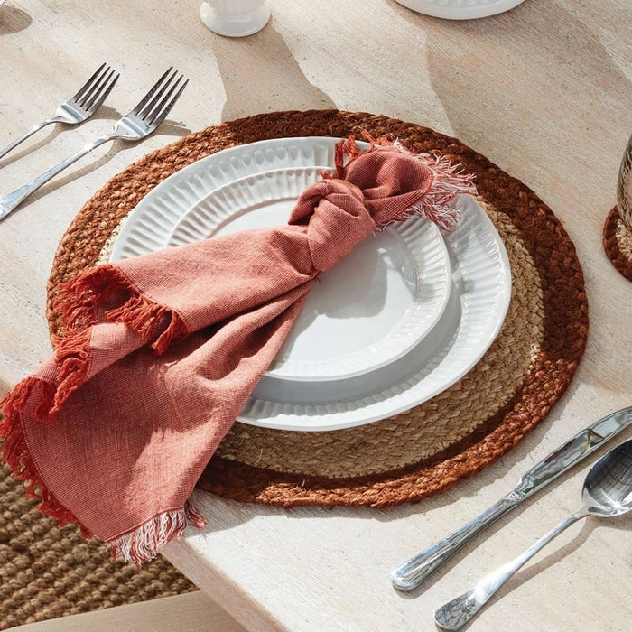 A table setting features white plates on woven placemats, a NAPA HOME AND GARDEN Aura Dinner Napkin in Rust with fringed edges knotted on top, and neatly arranged silverware on a light table.