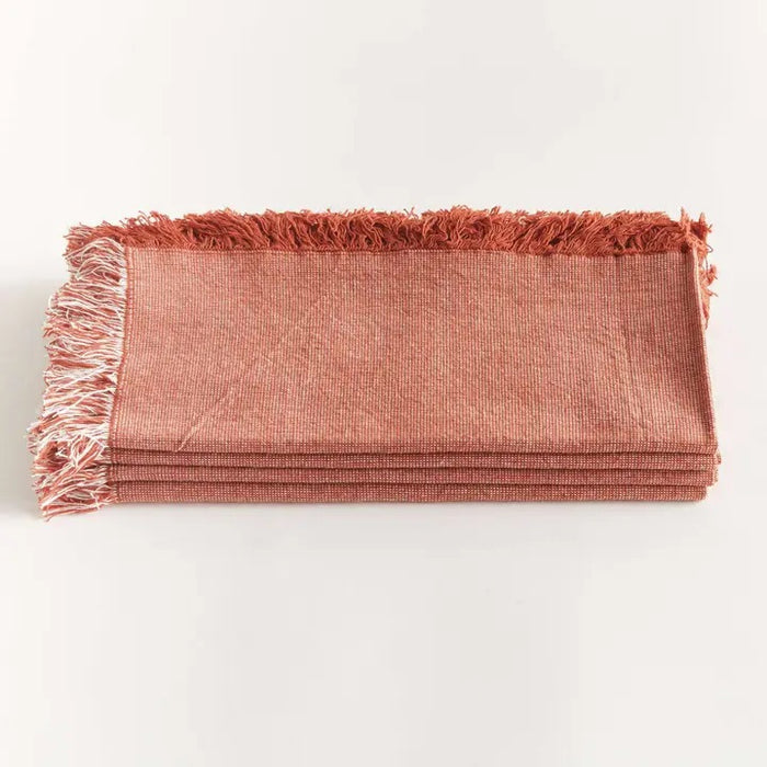 A table setting features white plates on woven placemats, a NAPA HOME AND GARDEN Aura Dinner Napkin in Rust with fringed edges knotted on top, and neatly arranged silverware on a light table.