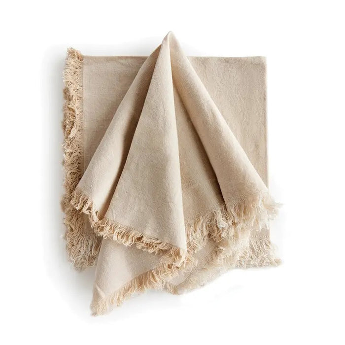 The NAPA HOME AND GARDEN - AURA Dinner Napkin in taupe is a square cotton napkin with fringed edges, neatly folded and set on a white background. Machine washable for easy care.