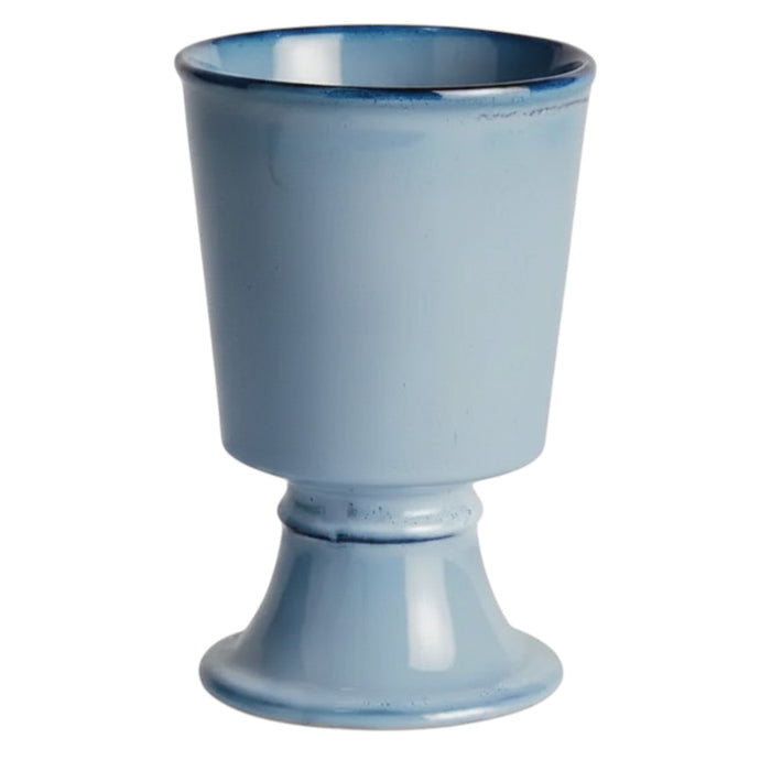 Two NAPA HOME AND GARDEN - BLUE KELCY FOOTED CACHEPOTS, one with a leafy plant and one empty, rest on a wooden table with a blue and white striped placemat, with a white lattice and wooden chair in the background.