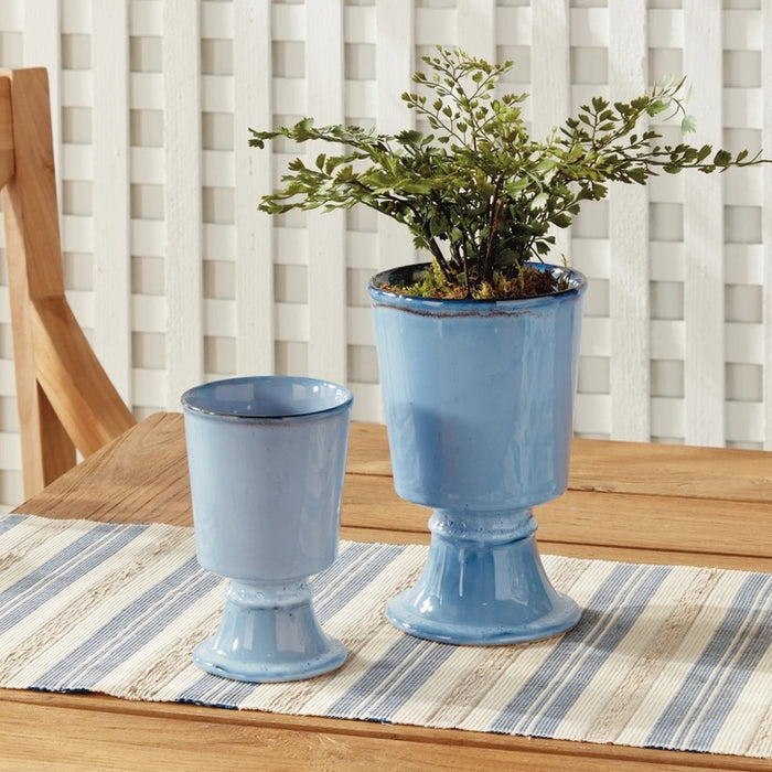 Two NAPA HOME AND GARDEN - BLUE KELCY FOOTED CACHEPOTS, one with a leafy plant and one empty, rest on a wooden table with a blue and white striped placemat, with a white lattice and wooden chair in the background.
