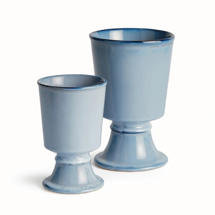 Two NAPA HOME AND GARDEN - BLUE KELCY FOOTED CACHEPOTS, one with a leafy plant and one empty, rest on a wooden table with a blue and white striped placemat, with a white lattice and wooden chair in the background.