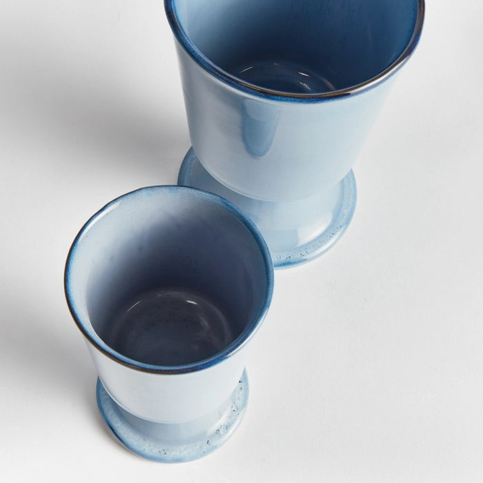 Two NAPA HOME AND GARDEN - BLUE KELCY FOOTED CACHEPOTS, one with a leafy plant and one empty, rest on a wooden table with a blue and white striped placemat, with a white lattice and wooden chair in the background.