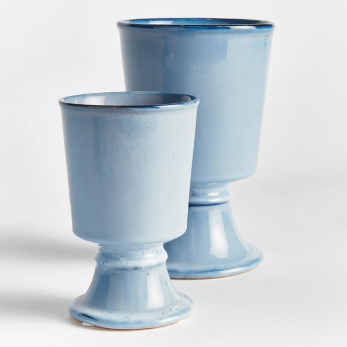 Two NAPA HOME AND GARDEN - BLUE KELCY FOOTED CACHEPOTS, one with a leafy plant and one empty, rest on a wooden table with a blue and white striped placemat, with a white lattice and wooden chair in the background.
