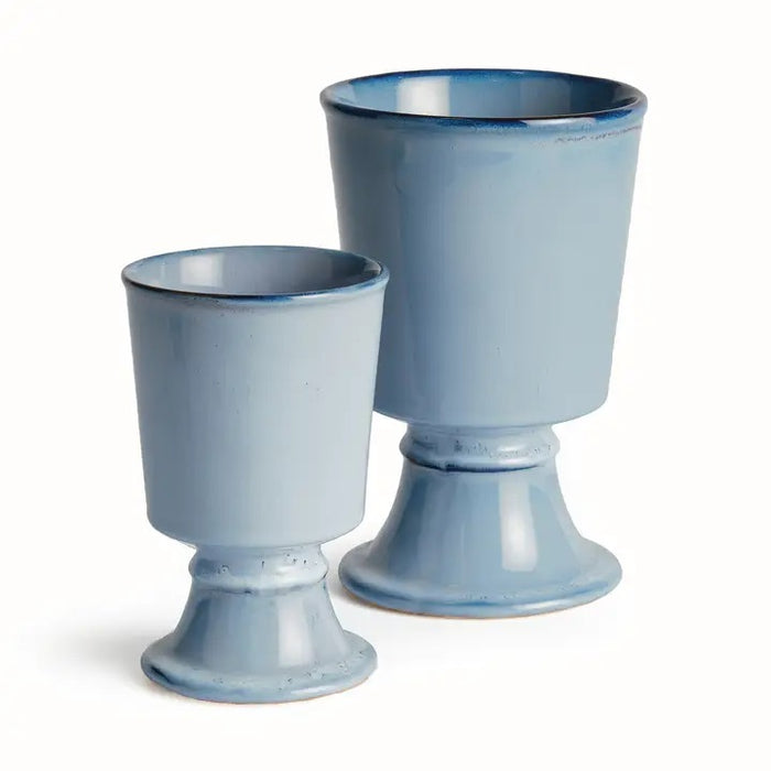 Two NAPA HOME AND GARDEN Blue Kelcy Footed Cachepot planters sit on a wooden table with a blue and white striped placemat; one holds a small leafy green plant, the other is empty. A white textured wall is in the background.