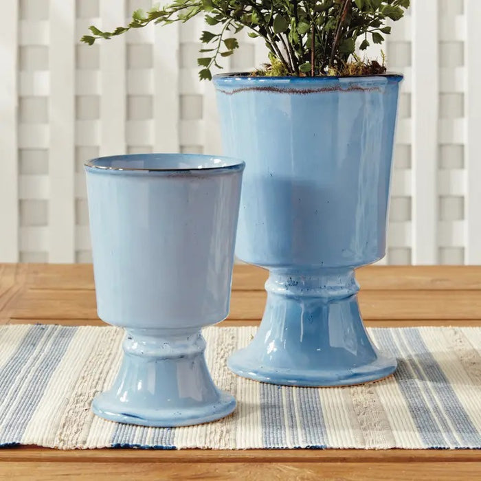 The NAPA HOME AND GARDEN Blue Kelcy Footed Cachepot, Small features a light blue ceramic finish with a glossy reactive glaze, subtle darker speckles near the rim and base, plus a wide rim and sturdy flared foot for a classic look.