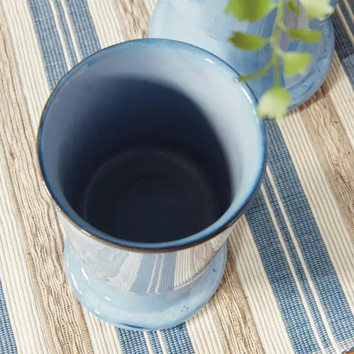Two NAPA HOME AND GARDEN Blue Kelcy Footed Cachepot planters sit on a wooden table with a blue and white striped placemat; one holds a small leafy green plant, the other is empty. A white textured wall is in the background.