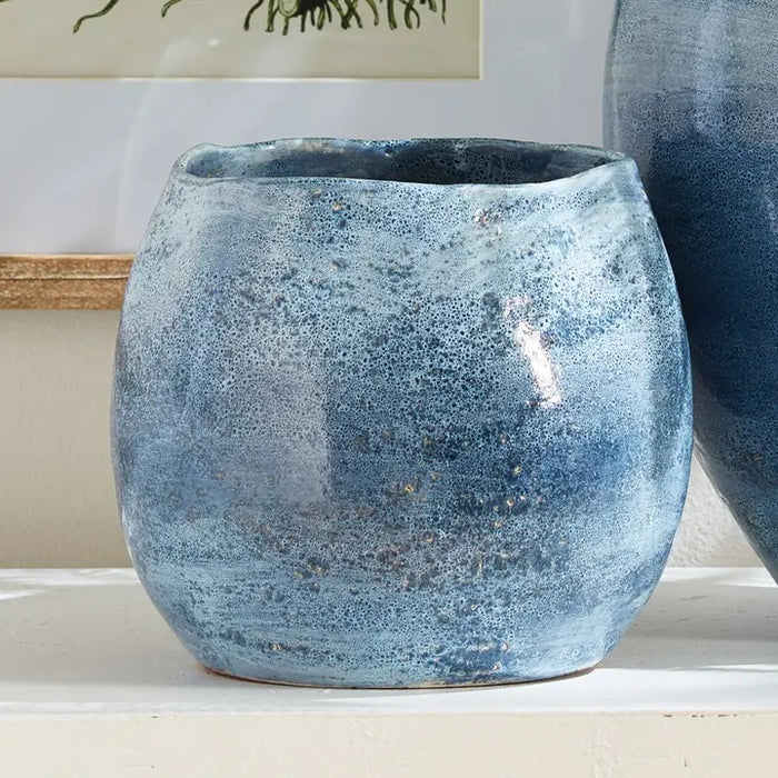 The NAPA HOME AND GARDEN - CASPIAN CACHEPOT is a round, blue ceramic vase with a glossy, mottled finish and speckled pattern—ideal for decorative use on any shelf or table.