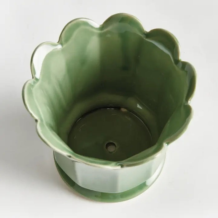 The NAPA HOME AND GARDEN - Maline Footed Pot in Green (Small) is a light green ceramic pot featuring scalloped edges and vertical panels, perched on a small round base, set against a plain white background.