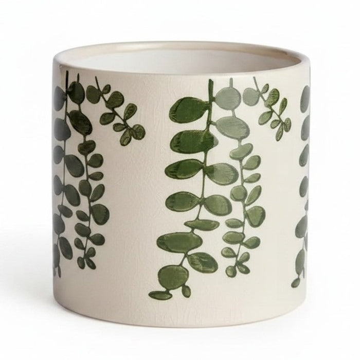 Two NAPA HOME AND GARDEN Maren Pots in white ceramic with an embossed leaf pattern sit on a wooden table, one with a leafy plant. Nearby are two stacked books and a partially visible empty green cachepot.