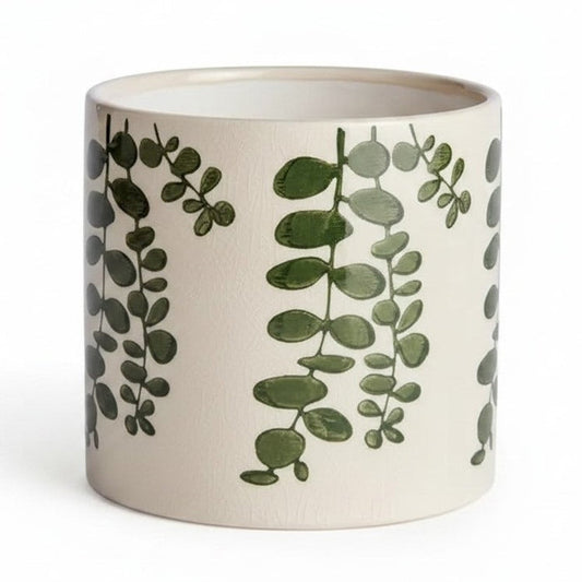 Two NAPA HOME AND GARDEN Maren Pots in white ceramic with an embossed leaf pattern sit on a wooden table, one with a leafy plant. Nearby are two stacked books and a partially visible empty green cachepot.