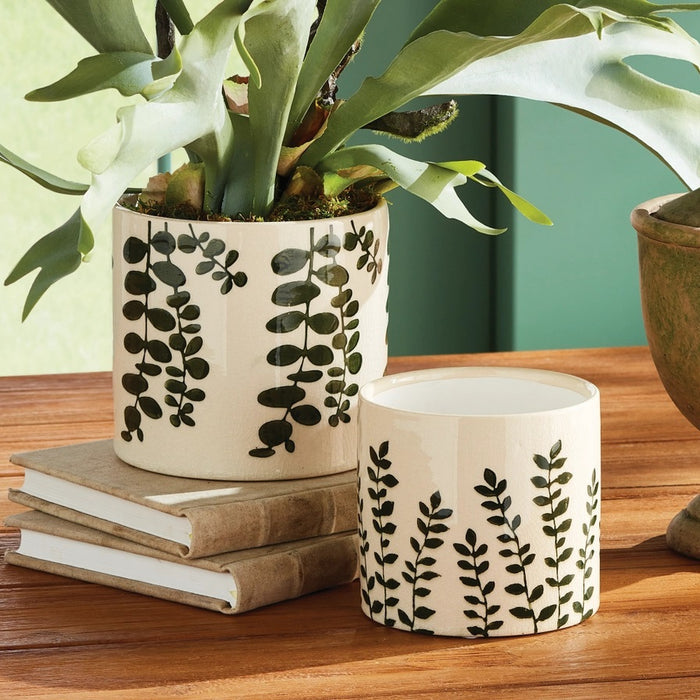 Two NAPA HOME AND GARDEN Maren Pots in white ceramic with an embossed leaf pattern sit on a wooden table, one with a leafy plant. Nearby are two stacked books and a partially visible empty green cachepot.