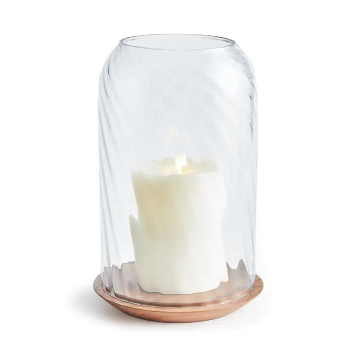 The NAPA HOME AND GARDEN - NIRA HURRICANE, LARGE features a white sculpted candle with a gold accent beneath ribbed blown glass on an acacia wood base, creating a striking decorative accent against a white background.