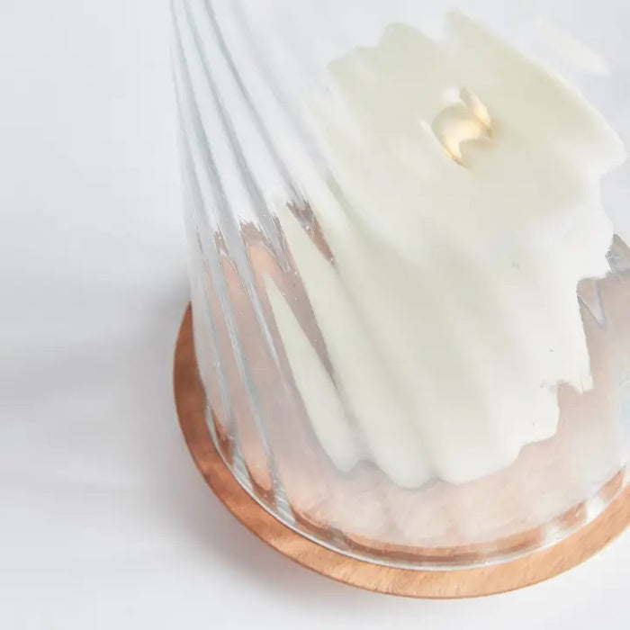 The NAPA HOME AND GARDEN - NIRA HURRICANE, LARGE features a white sculpted candle with a gold accent beneath ribbed blown glass on an acacia wood base, creating a striking decorative accent against a white background.