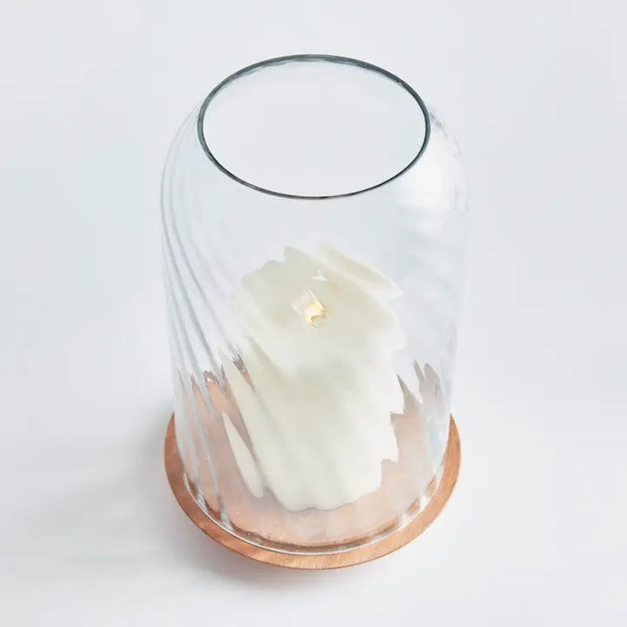 The NAPA HOME AND GARDEN - NIRA HURRICANE, LARGE features a white sculpted candle with a gold accent beneath ribbed blown glass on an acacia wood base, creating a striking decorative accent against a white background.