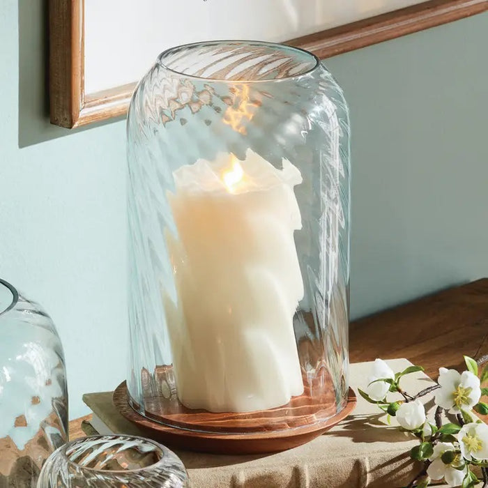 The NAPA HOME AND GARDEN - NIRA HURRICANE, LARGE features a white sculpted candle with a gold accent beneath ribbed blown glass on an acacia wood base, creating a striking decorative accent against a white background.