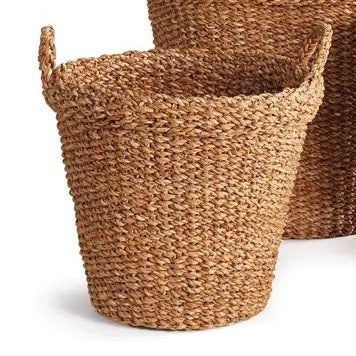 The NAPA HOME AND GARDEN Seagrass Tapered Basket with Handles and Cuff, Small, sits on a textured rug before a white paneled wall, alongside two other baskets, with part of a chair and a colorful painting in the background.