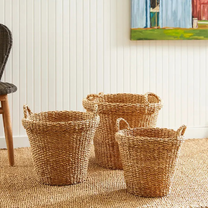 The NAPA HOME AND GARDEN Seagrass Tapered Basket with Handles and Cuff, Small, sits on a textured rug before a white paneled wall, alongside two other baskets, with part of a chair and a colorful painting in the background.