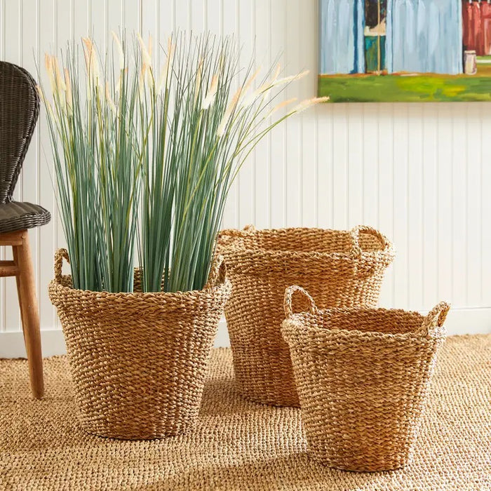 The NAPA HOME AND GARDEN Seagrass Tapered Basket with Handles and Cuff, Small, sits on a textured rug before a white paneled wall, alongside two other baskets, with part of a chair and a colorful painting in the background.